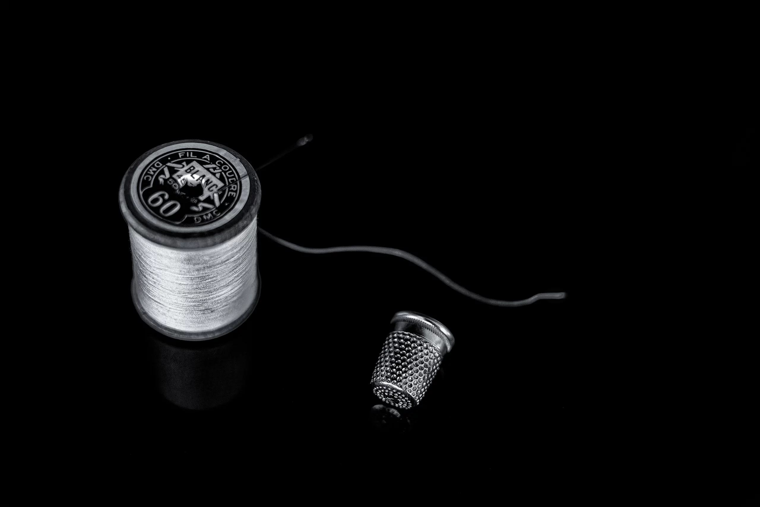 Photographie en noir et blanc d’une bobine de fil et d’un dé à coudre posés sur un fond noir, éclairés de façon à faire ressortir les textures du métal et du fil.