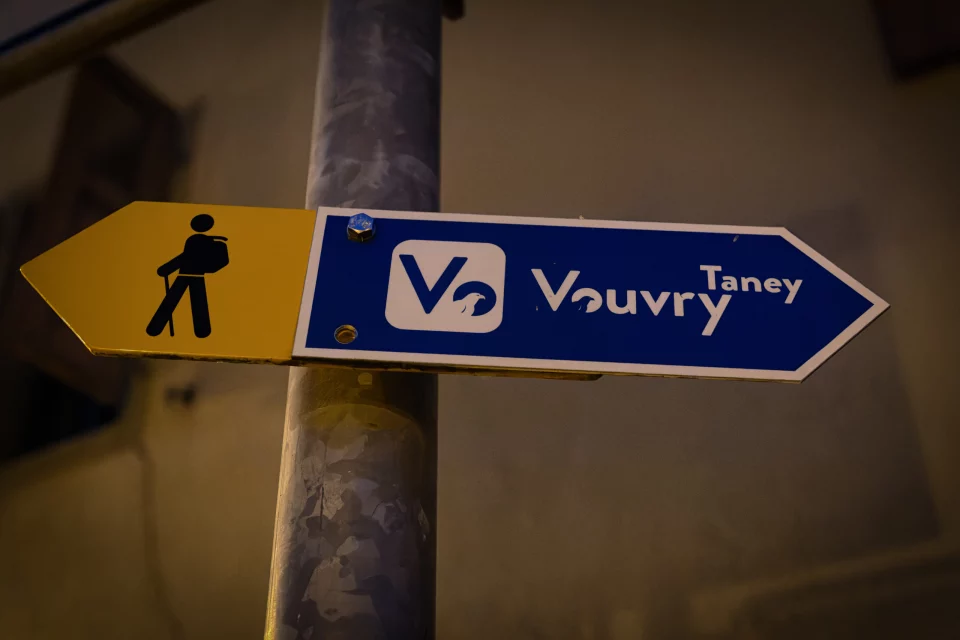Panneau directionnel bleu et jaune indiquant Vouvry et Taney, photographié de nuit.