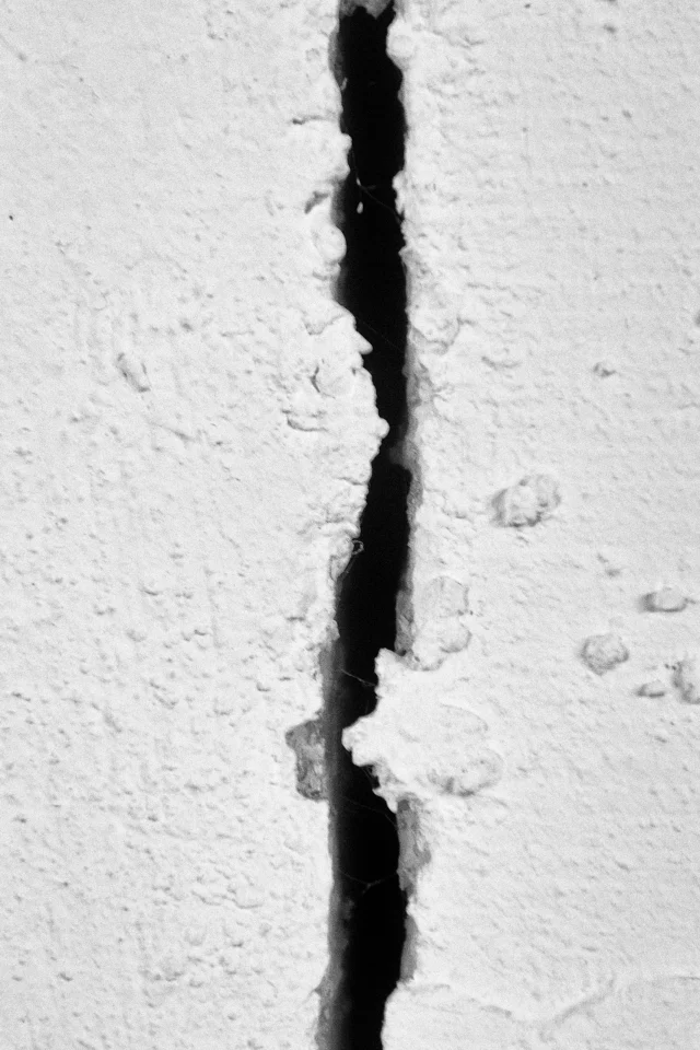 Gros plan en noir et blanc d’une fissure verticale sombre traversant un mur clair à la surface rugueuse.
