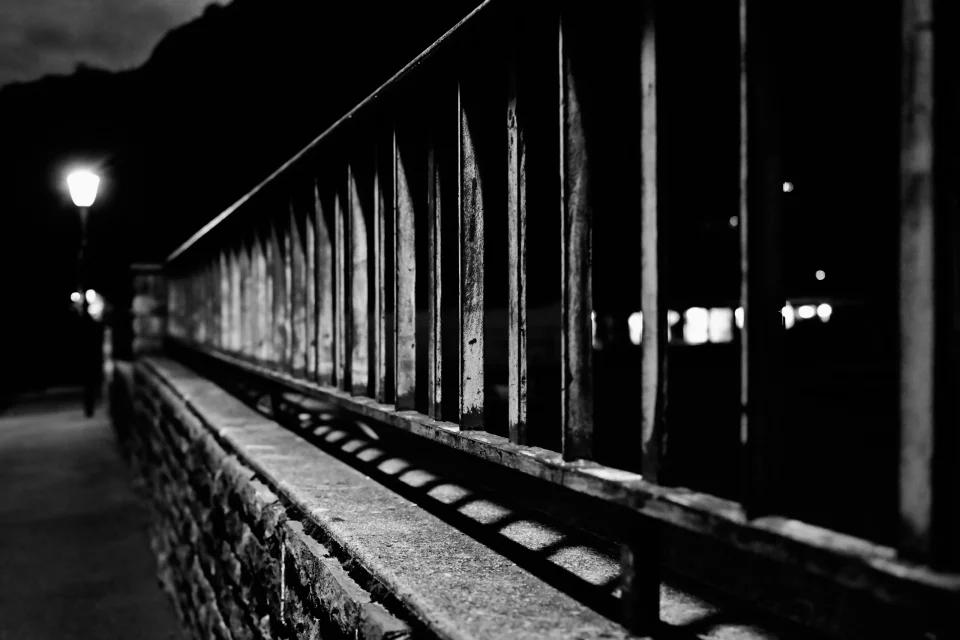 Photographie noir et blanc nocturne d’une rambarde métallique longeant un muret, éclairée par un lampadaire, avec une forte perspective et des ombres géométriques.
